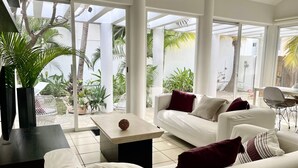 Ein modernes Wohnzimmer mit einem weißen Sofa, einem Couchtisch und einem Fernseher. Große Fenster bieten Blick auf einen Garten und Palmen.