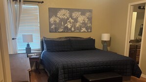 Quarto com uma cama grande, mesa de cabeceira, ventilador de teto, janela com persianas e um quadro pendurado na parede.