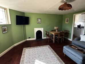 Un salon avec une cheminée, une table à manger avec des chaises, un téléviseur fixé au mur et un coin cuisine visible à travers une porte ouverte.