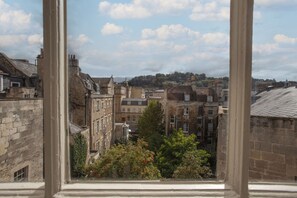 Une vue à travers une fenêtre sur un paysage urbain, avec des bâtiments en pierre, une colline au loin et un ciel dégagé.