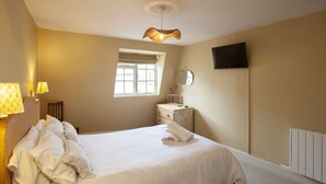A bedroom with a bed, bedside table, lamp, window, mirror, and a flat-screen TV.