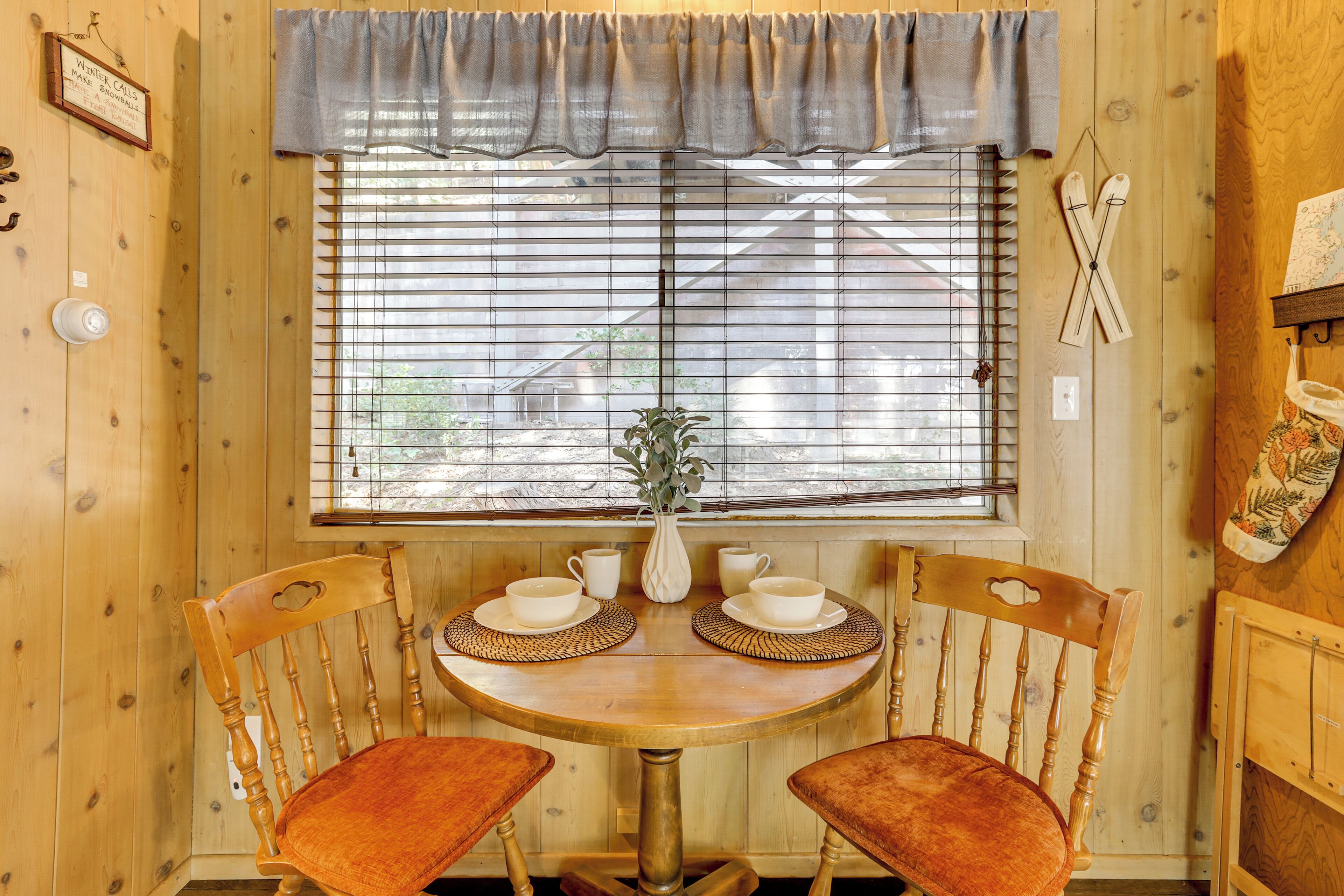 Un interior de madera con una mesa redonda para dos, una ventana con persianas y un perchero en la pared.