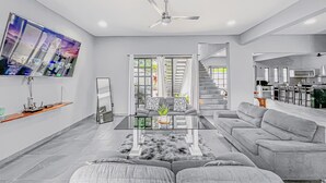A modern living room with a flat-screen TV, a ceiling fan, and a staircase leading to a kitchen area.