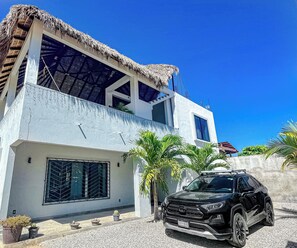 A modern two-story house with a thatched roof, a black SUV parked in front, and a clear blue sky.