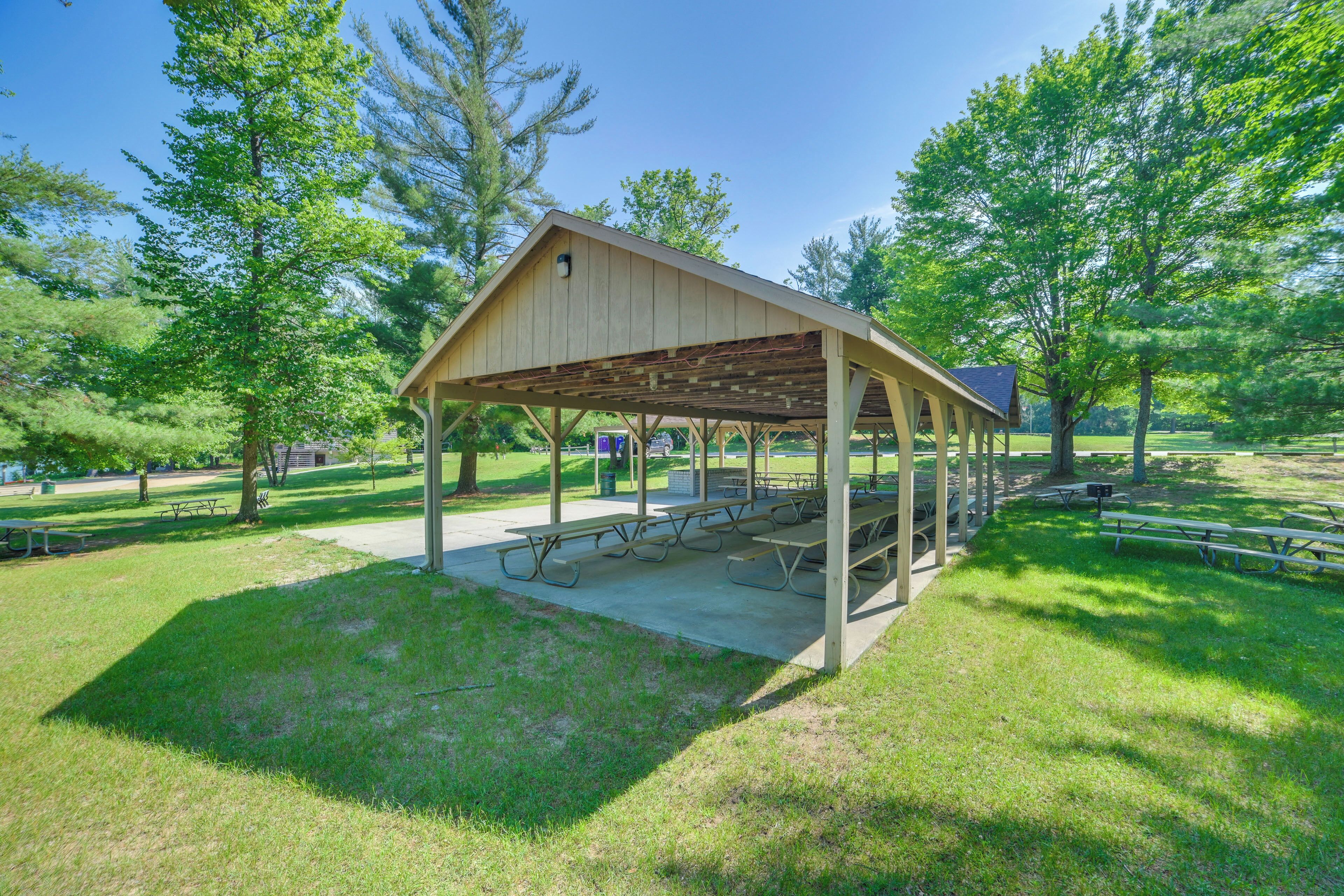 A covered picnic area with benches and picnic tables, surrounded by trees and grass.
