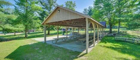 A covered picnic area with benches and picnic tables, surrounded by trees and grass.
