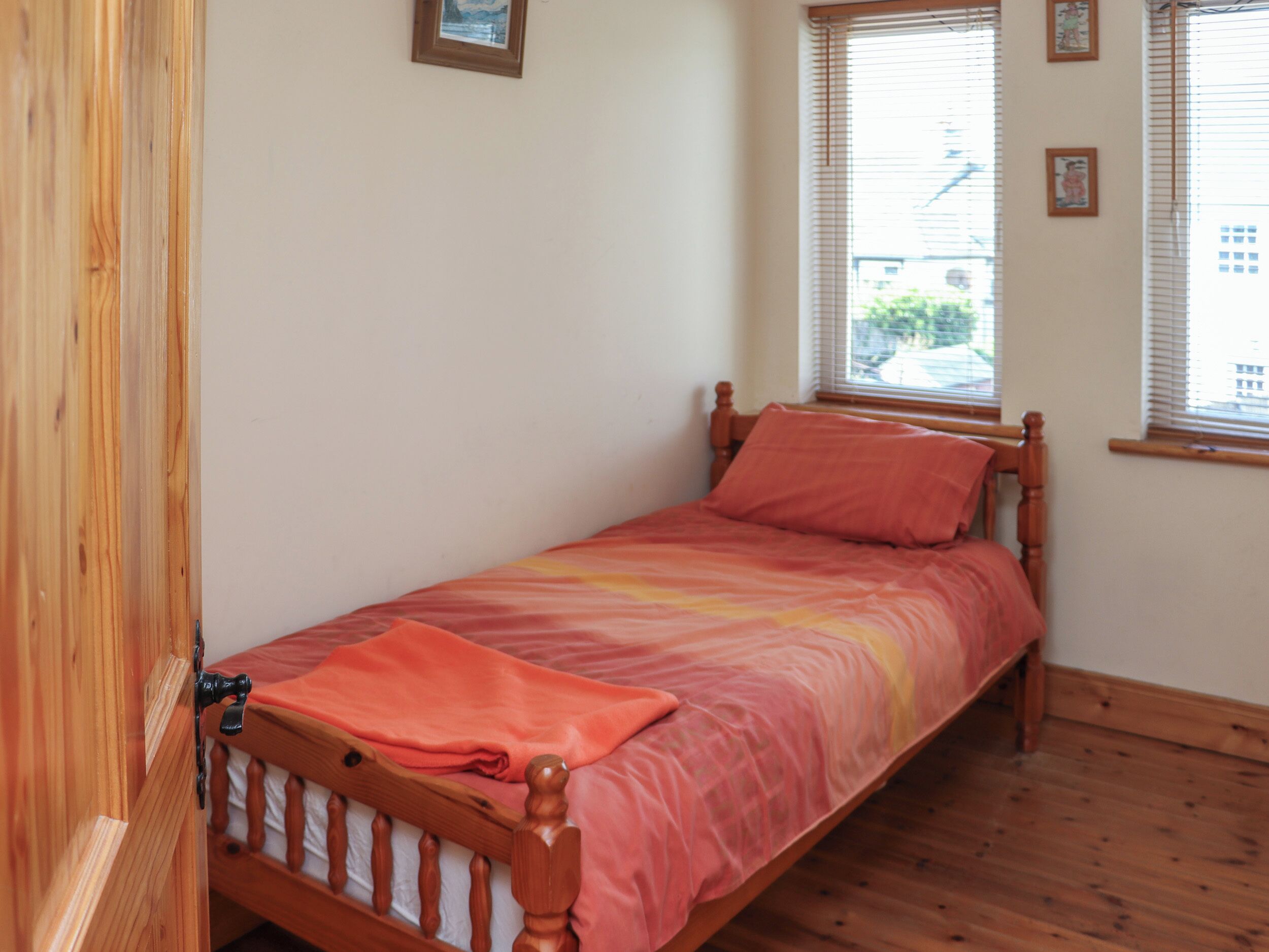 Un lit simple avec un cadre en bois et une couvre-lit rose, placé contre un mur où se trouvent une fenêtre et une porte.