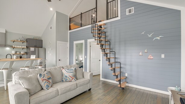 A spacious living area with a grey sofa, wooden staircase, and a kitchen in the background.