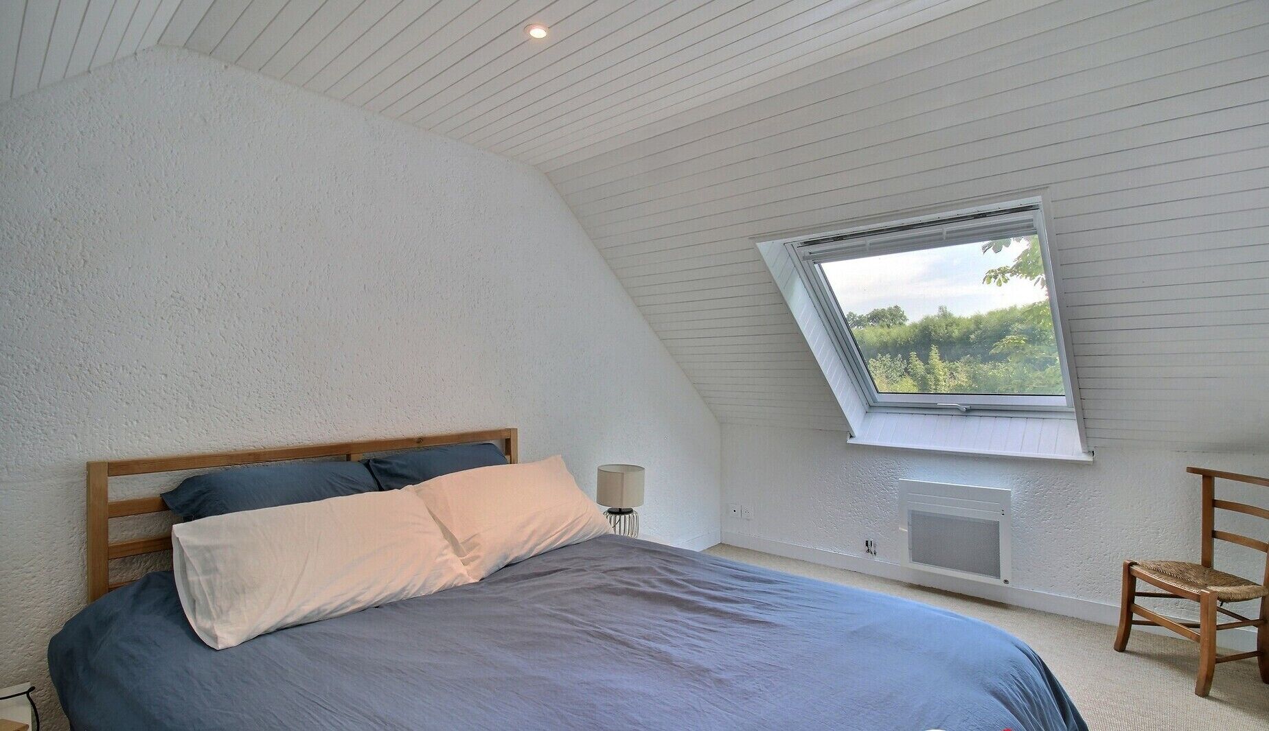 A bedroom with a sloped ceiling, a skylight, a bed with blue bedding, a bedside lamp, and a chair.