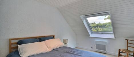A bedroom with a sloped ceiling, a skylight, a bed with blue bedding, a bedside lamp, and a chair.