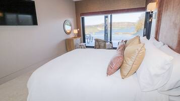 Ein großes Bett mit vielen Kissen, ein Nachttisch mit Lampe, ein an der Wand befestigtes Fernsehgerät und ein Blick auf einen See durch ein großes Fenster.