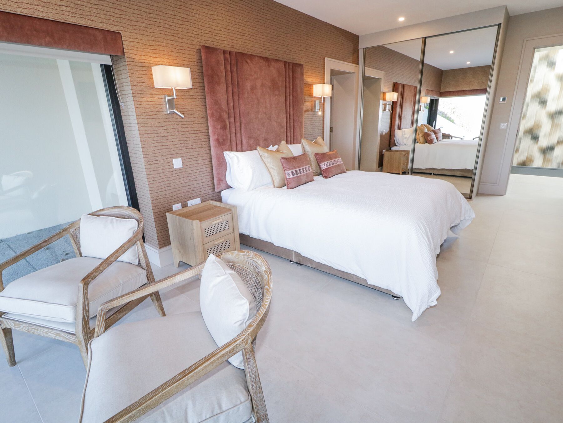 A modern hotel room with a large bed, two bedside tables, a small wooden nightstand, a wicker chair, and a large mirror.