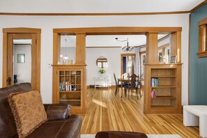 Una sala de estar con un sofá de cuero marrón, estanterías de madera, un comedor con candelabro y suelo de madera.