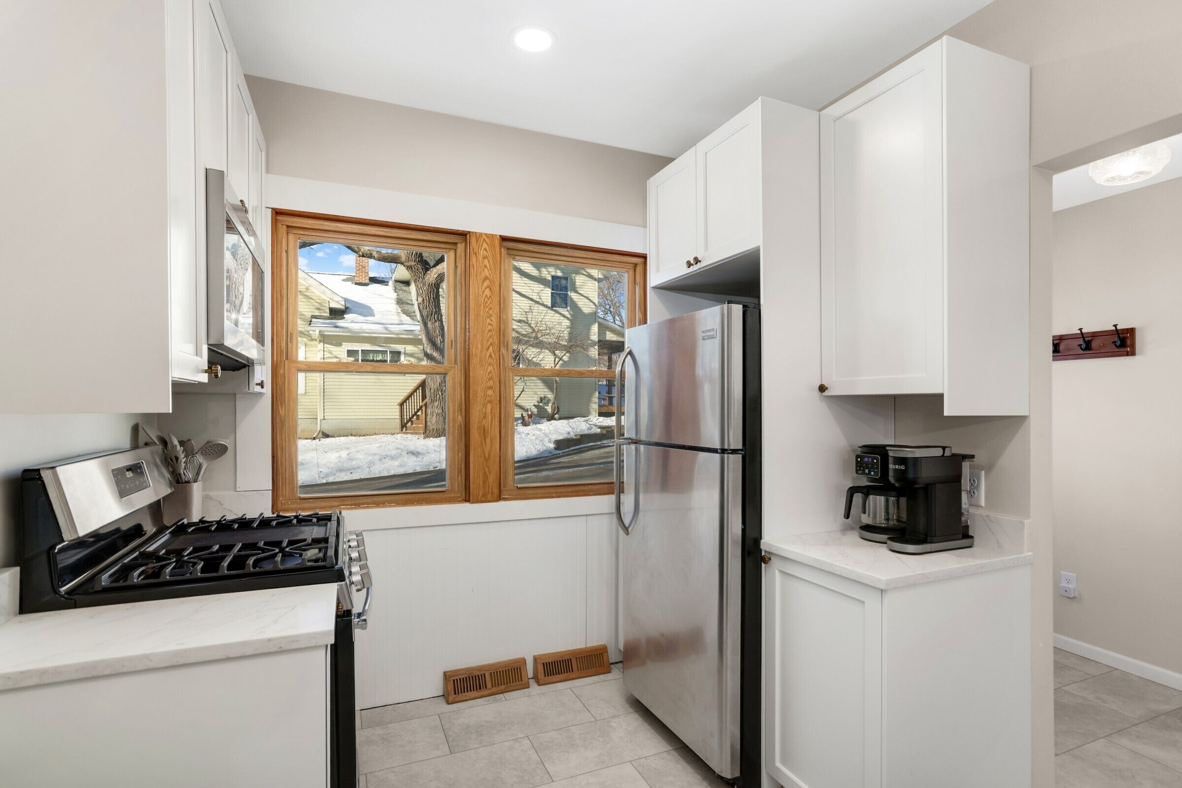 A compact kitchen with white cabinets, a stainless steel refrigerator, a coffee maker, and a window with a view of a snowy yard.