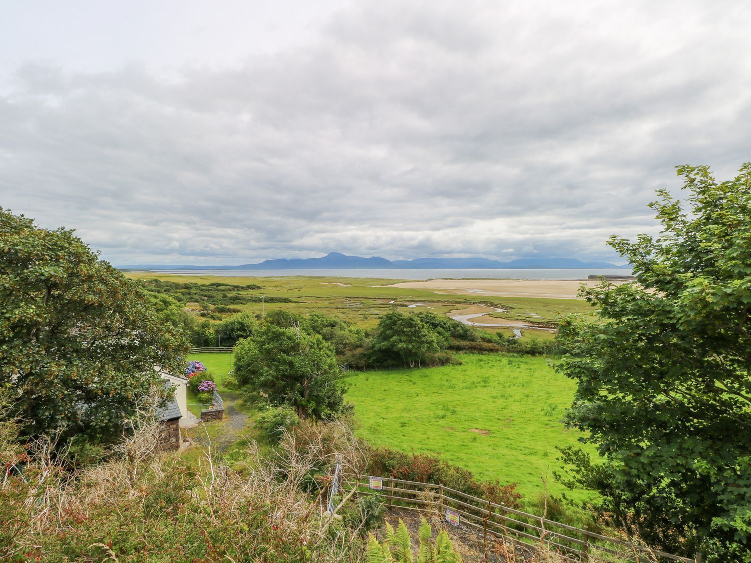 A rural landscape with a view of a grassy field, a small building, and distant mountains under a cloudy sky.