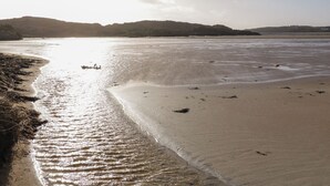 A wide sandy beach with gentle waves, a clear sky, and distant hills.