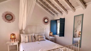 Un dormitorio con cama a dosel, dos mesitas de noche con lámparas, un ventanal con contraventanas azules y un espejo en la pared.