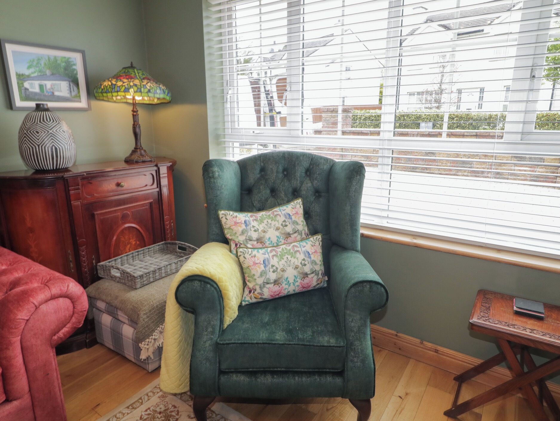 A cozy room with a green armchair, a wooden dresser, a lamp, and a window with blinds.