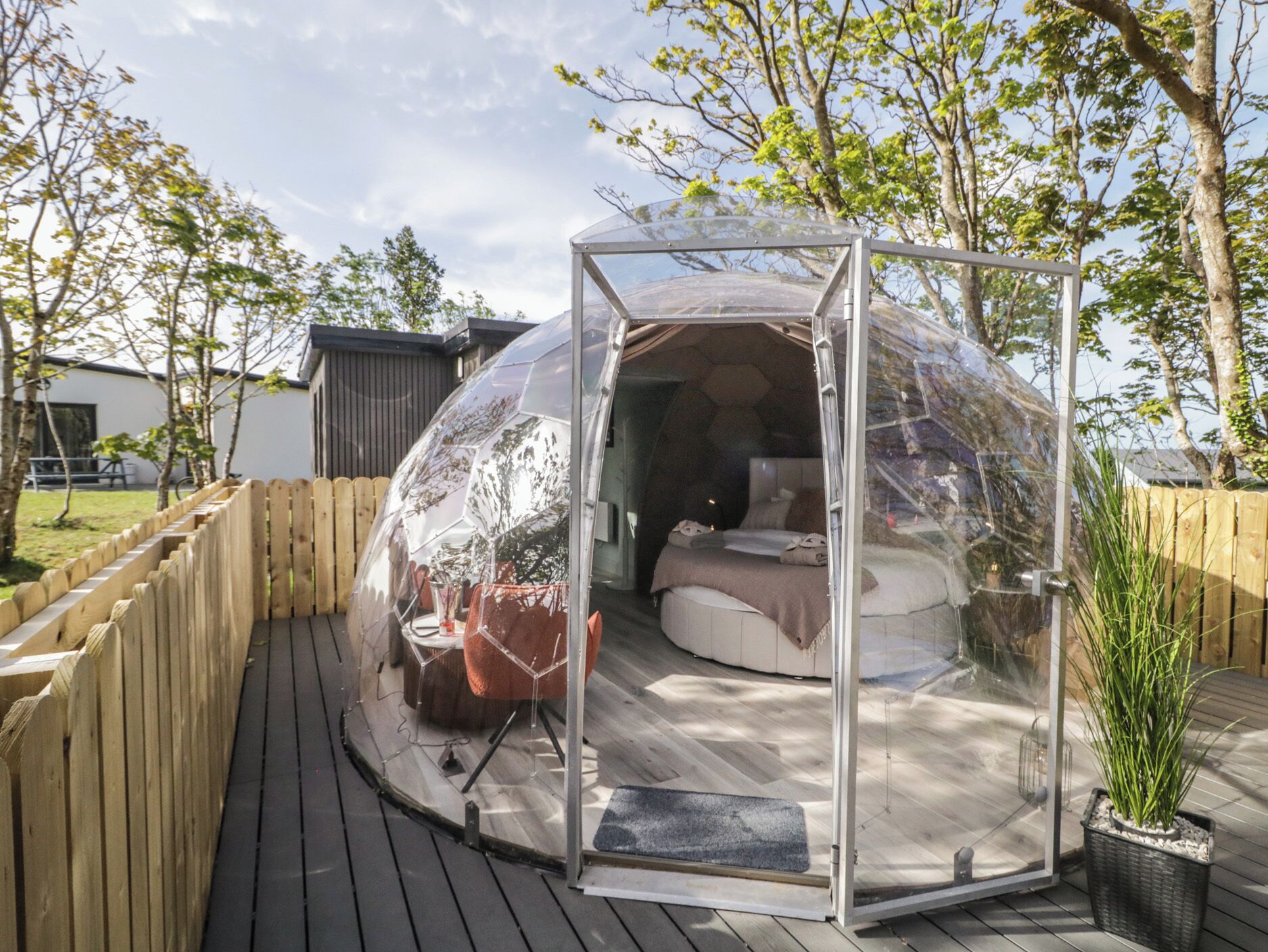 A transparent dome structure with a bed and seating area inside, surrounded by a wooden deck and a fence.
