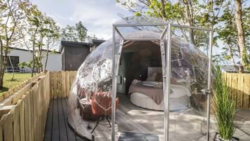 A transparent dome structure with a bed and seating area inside, surrounded by a wooden deck and a fence.
