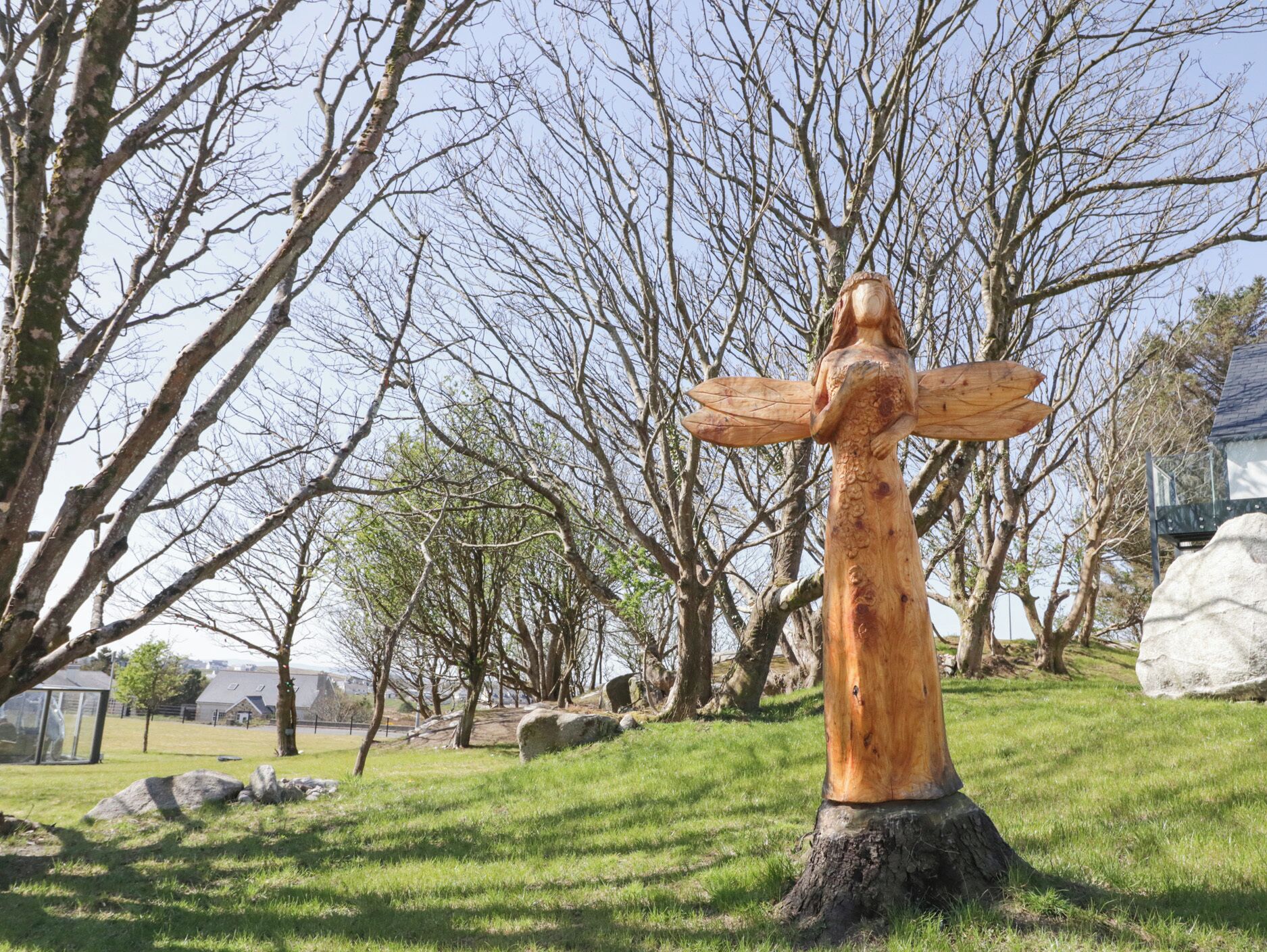 Escultura de fusta d'una dona amb els braços estesos a un parc amb arbres i roques.