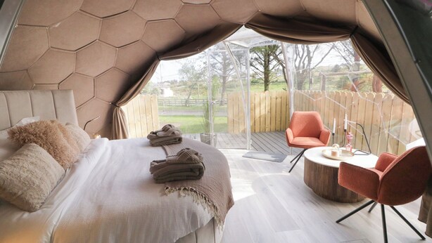 Ein Zelt-Schlafzimmer mit Bett, zwei Stühlen, einem kleinen Tisch und Blick auf eine Holzterrasse und Bäume.