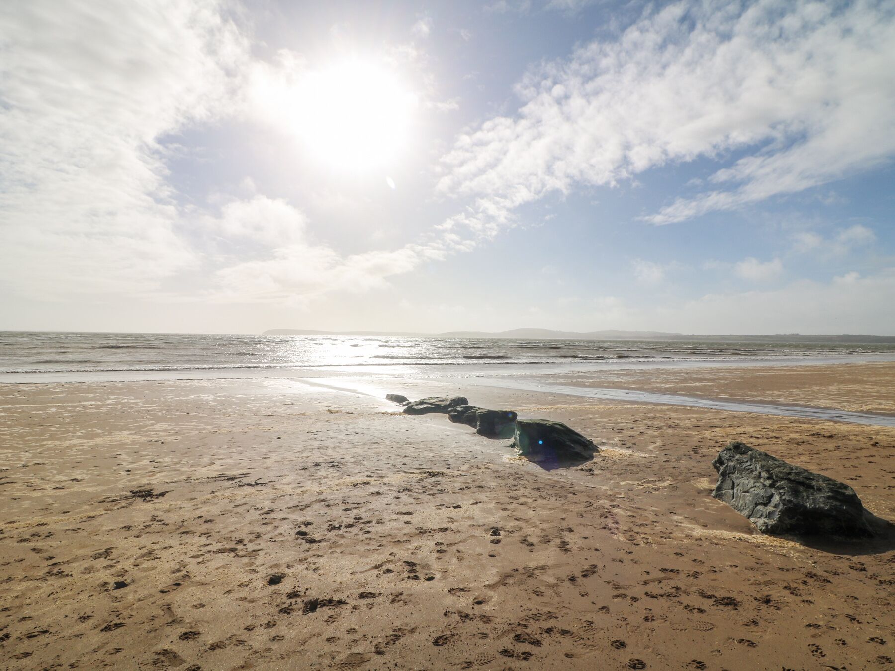 A beach with footprints and rocks, and a clear sky with the sun shining.