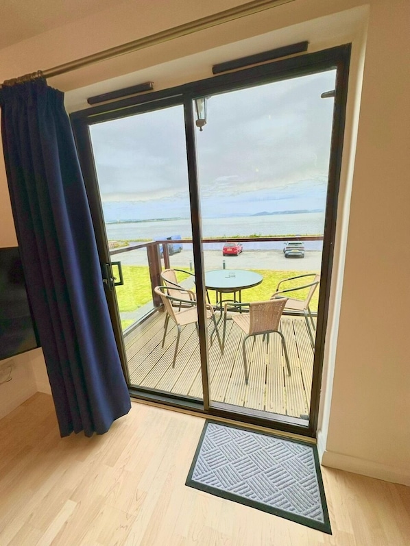 Ein Zimmer mit einer GlasschiebetĂŒr, die zu einem AuĂenbereich mit Tisch und StĂŒhlen, einer FuĂmatte und Blick auf das Meer sowie geparkte Autos fĂŒhrt.