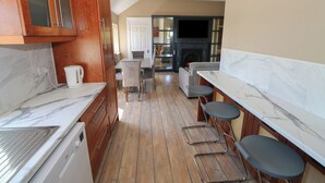 A modern kitchen with wooden cabinets, a marble countertop, and a built-in oven. There are two bar stools and a dining table with chairs. A fireplace is visible in the background.