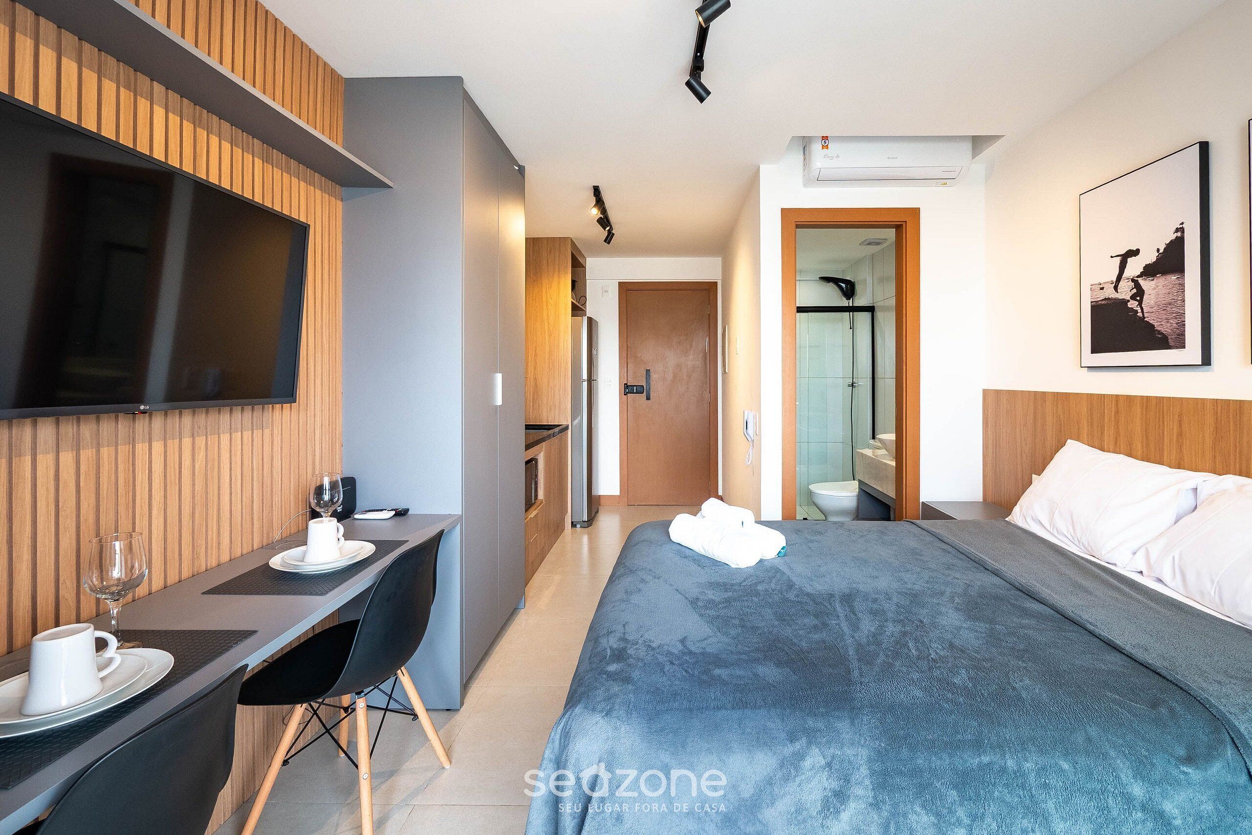 A hotel room with a bed, a desk with a chair, a TV mounted on the wall, and a bathroom visible through an open door.
