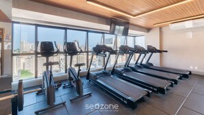 A gym with treadmills, stationary bikes, and a large window with a city view. - Modern and Complete Apartments 500m from Barra Beach in Salvador – ZNB (Salvador)