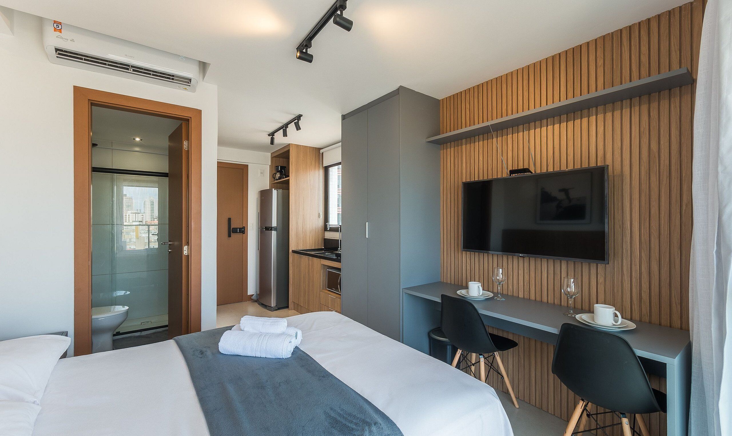 A modern hotel room with a bed, a desk, a TV, and a kitchenette.