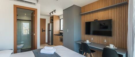 A modern hotel room with a bed, a desk, a TV, and a kitchenette.
