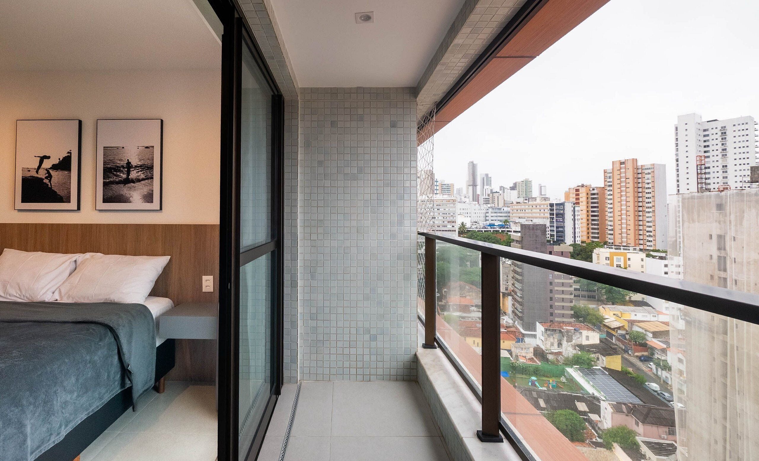 Un balcone con vista sulla città, un letto con coperta grigia e due quadri appesi al muro.