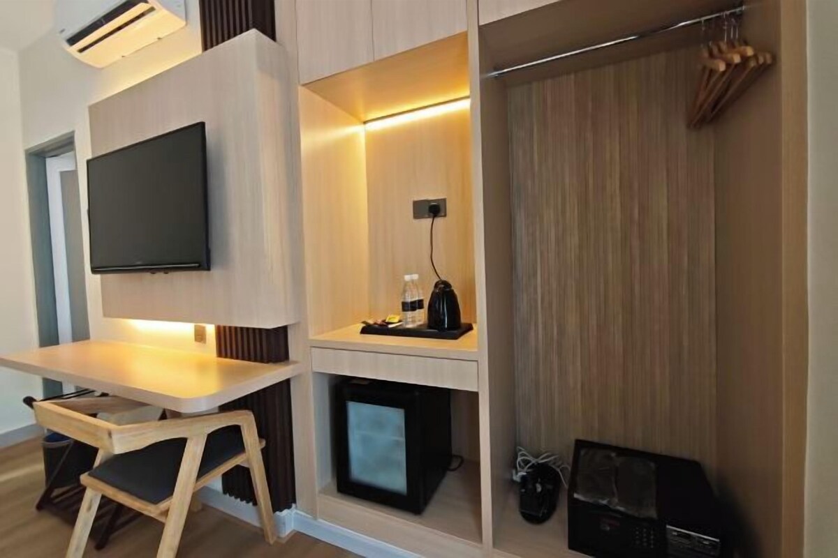 Habitación moderna con televisión de pantalla plana en la pared, una pequeña mesa con una silla, un refrigerador pequeño y un perchero con objetos colgados.
