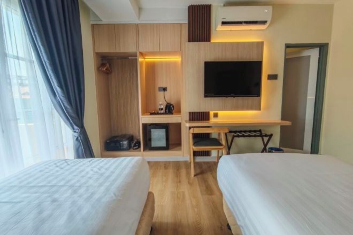 Quarto de hotel com duas camas, televisão, secretária com cadeira, guarda-roupa e ar condicionado.