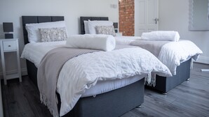 A modern bedroom with two beds, a white and gray color scheme, and a brick wall visible through an open door. - Hillview Modern 3 Bed Bromley, Chislehurst House (Greater London)
