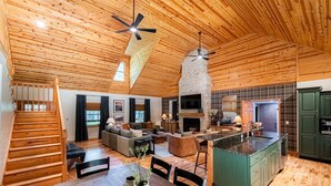Amplia sala de estar con techo de madera, chimenea de piedra y cocina con armarios verdes.