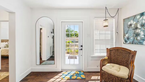 A clean, modern entryway with a wooden floor, a wicker chair, a door with a glass panel, and a large mirror.