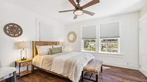 A bedroom with a wooden bed, a bench, a nightstand with a lamp, a ceiling fan, and a view of the outside through a window.