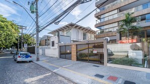 A street view with a gated entrance, a parked car, and a modern building with large windows. -  Apartamentos em Prédio c/Academia na Ondina-SSA-HOA (Salvador)