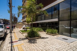A modern building with large glass windows, a paved sidewalk with tactile paving, and planters with green plants.