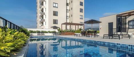 A swimming pool with tiled flooring, surrounded by lounge chairs and umbrellas, with a tall building in the background.