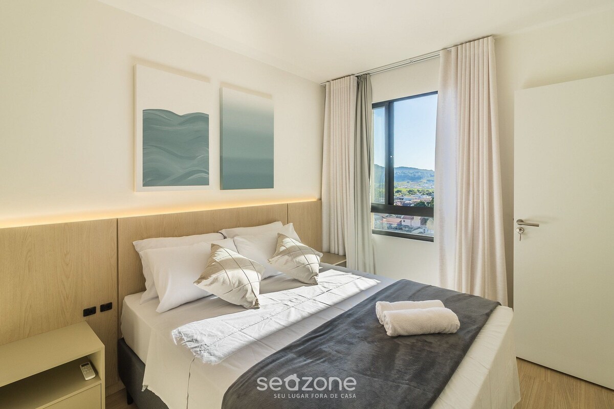 A hotel room with a bed, a nightstand, a window with curtains, and artwork on the wall.