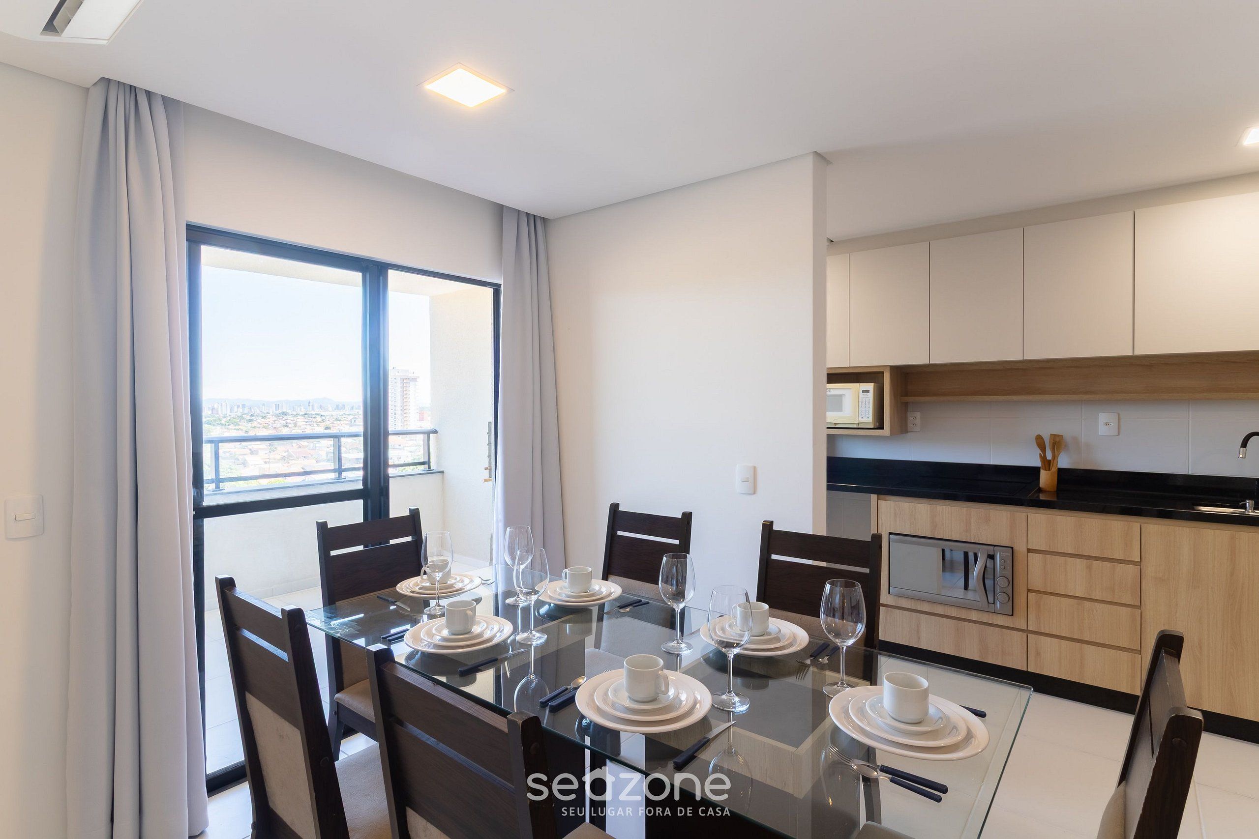 Uma sala de jantar moderna com uma mesa de vidro posta para seis pessoas, cadeiras de madeira e vista através de amplas janelas.