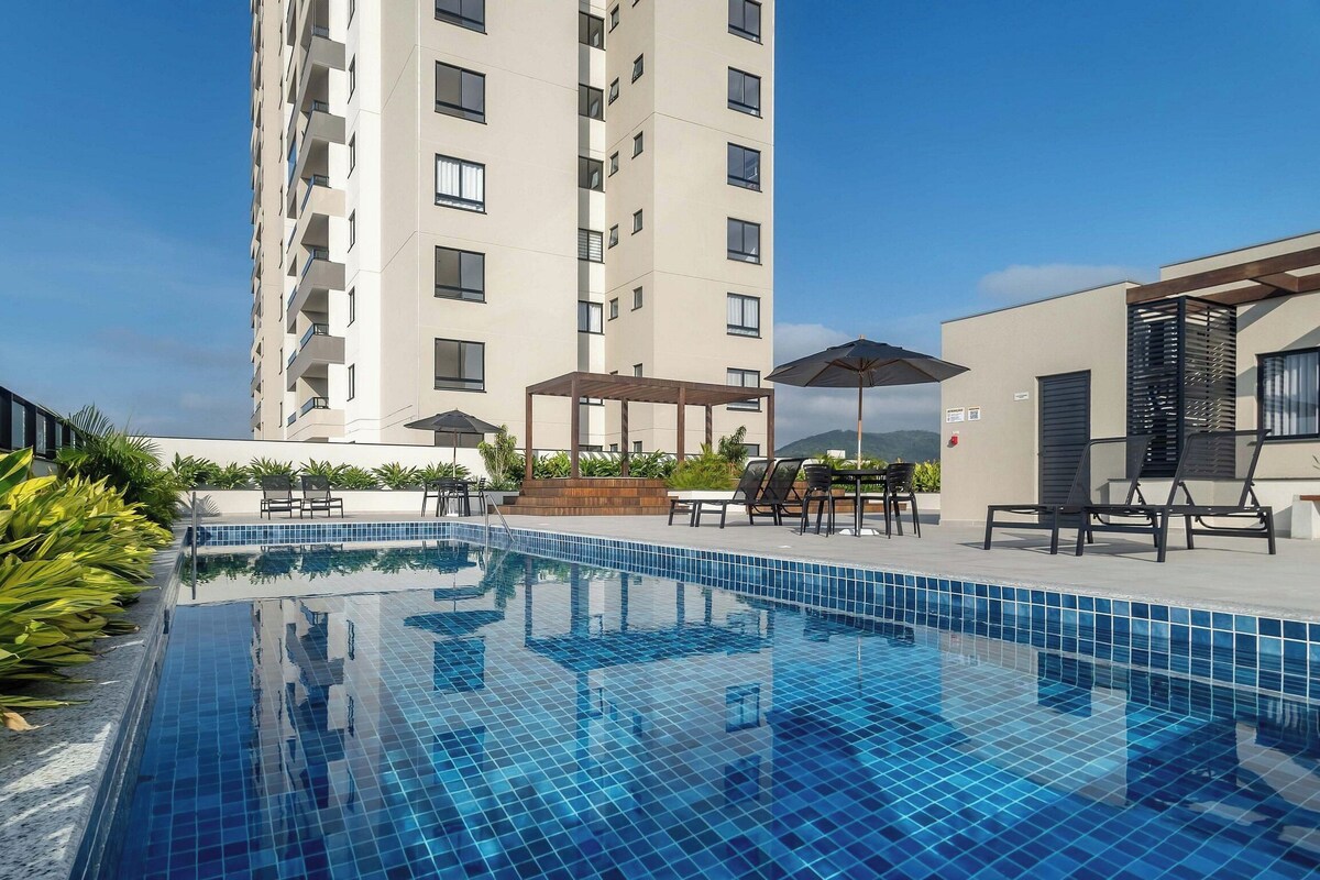 Une piscine dont le bord est revêtu de carrelage, entourée de chaises longues et de parasols, avec un grand immeuble en arrière-plan.