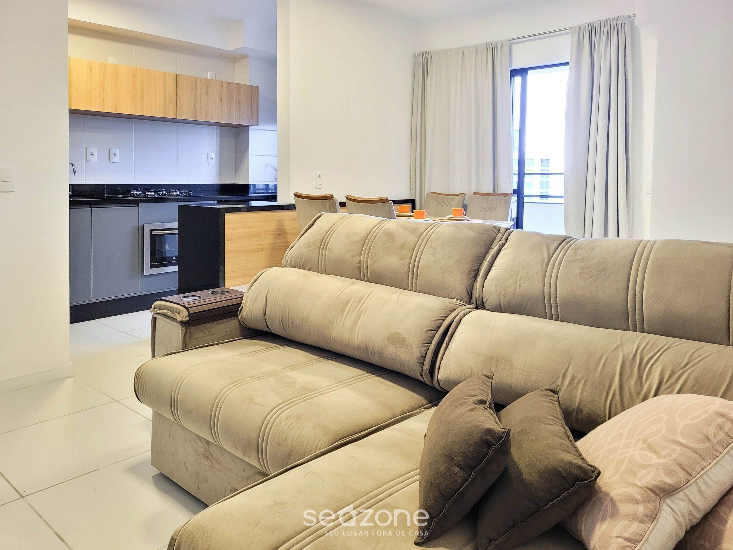 A modern living room with a beige sofa, a dining area with chairs, and a kitchen in the background.