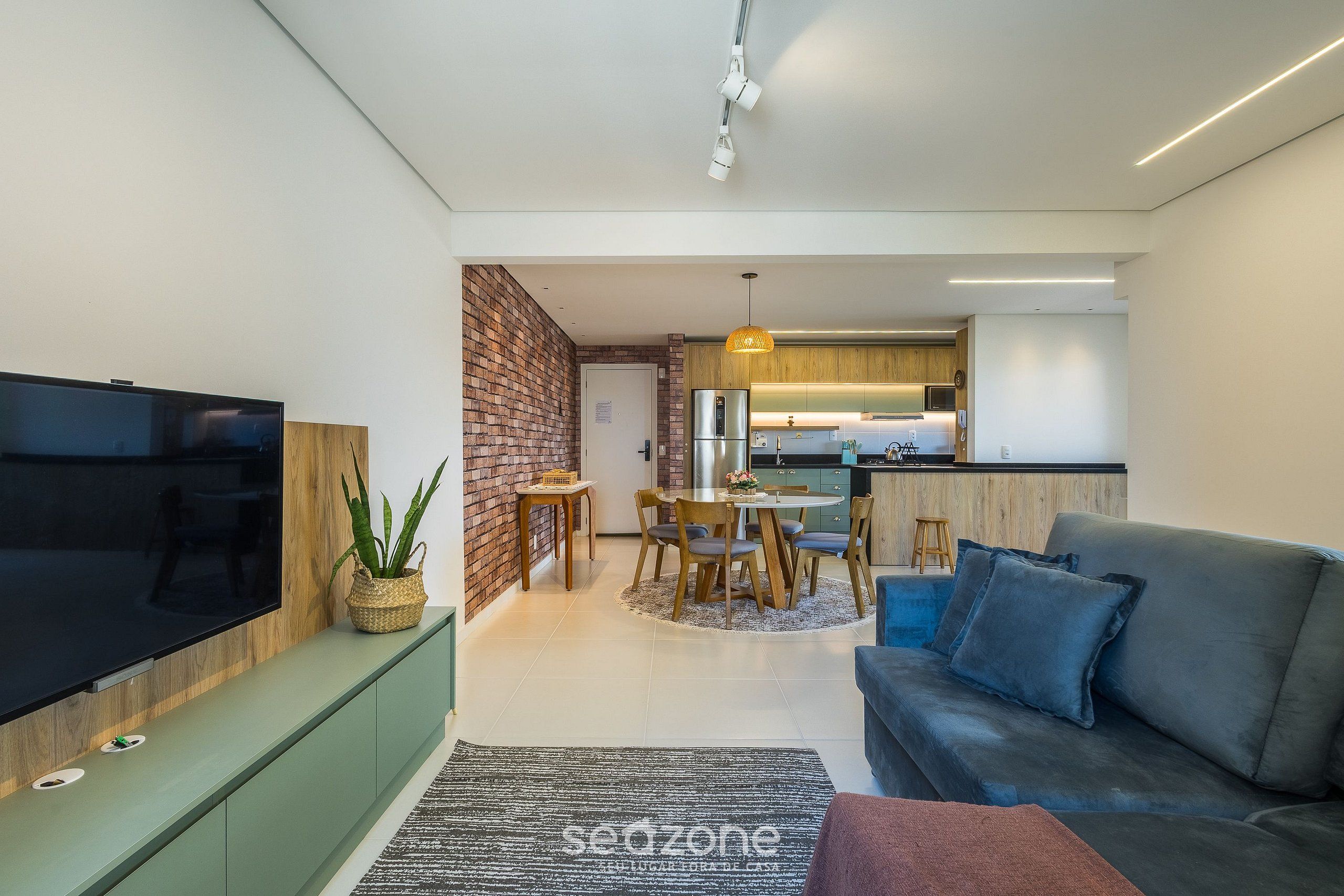 A modern living room with a flat-screen TV, a sofa, a dining area with a table and chairs, and a kitchen in the background.