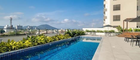 Una piscina en la azotea con suelo de baldosas, rodeada de vegetación y mobiliario de exterior.