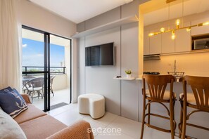 A modern living area with a sofa, ottoman, and wooden chairs, a TV mounted on the wall, and a balcony with a view.